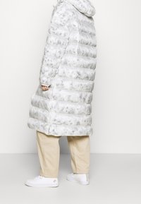 Person som bär en lång vit och gråmönstrad pufferjacka med beige byxor och vita sneakers, stående mot en enfärgad vit bakgrund.