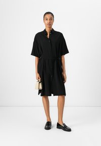 Black shirt dress with short sleeves, belt at waist, button closure, and side slits. Model wearing black loafers, holding a small beige handbag.