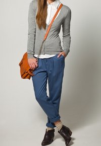 Grau gestrickte Jacke über einem weißen Hemd, kombiniert mit blauen Hosen. Braune Knöchelstiefel, orangefarbige Handtasche und hochgerollte Hosenbeine.