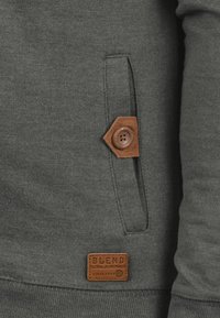 Szary sweatshirt z naszywaną kieszenią, wyposażoną w brązowy skórzany guzik i metkę, charakteryzujący się miękką fakturą i detalem ściągacza na dole.