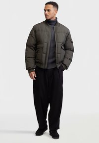 Geacă puf olive-verde cu guler striat, buzunare frontale și închidere cu fermoar, purtată peste un pulover tricotat închis la culoare și pantaloni negri lejeri.