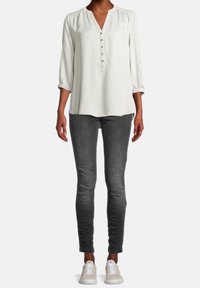Blusa grigia chiara a maniche lunghe con chiusura a bottone e dettaglio sulle spalle plissettato, abbinata a jeans slim-fit grigi scuri e sneakers chiare.