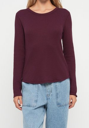 Vrouw draagt een donkerpaarse, ribgebreide top met lange mouwen, gecombineerd met lichtblauwe, hoogtailerende spijkerbroek met grote voorzakken.