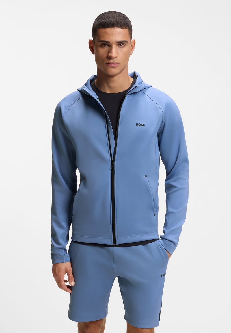 Νεαρός άνδρας που φοράει ανοιχτό μπλε φούτερ με φερμουάρ και παντελόνια σορτς που ταιριάζουν, με μαύρο εσώρουχο, κοιτώντας μπροστά με φόντο απλό.