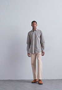 Hombre de pie contra una pared gris claro uniforme, vistiendo una camisa de cuadros de manga larga, pantalones beiges y zapatos marrones.