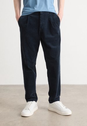 Personne portant un pantalon en velours côtelé bleu marine et des baskets blanches, debout les mains dans les poches devant un fond uni.