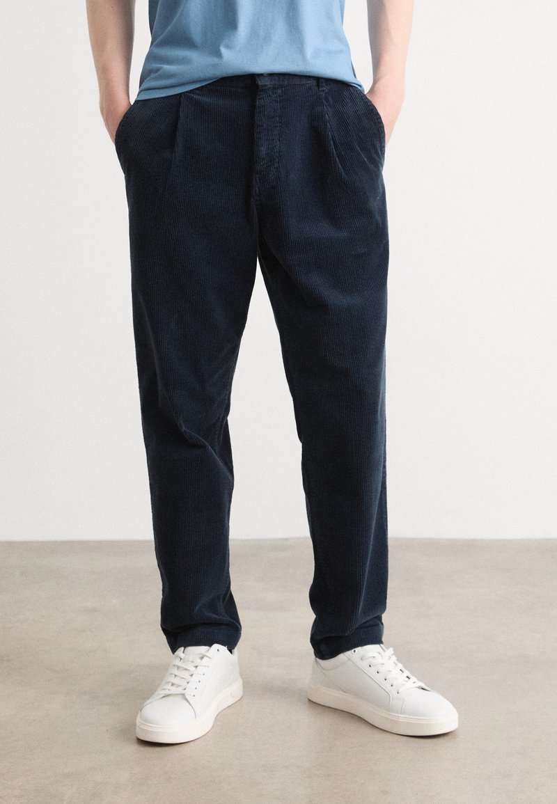 Personne portant un pantalon en velours côtelé bleu marine et des baskets blanches, debout les mains dans les poches devant un fond uni.