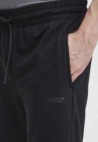 Schwarze Sporthose aus weichem Material, ausgestattet mit einem elastischen Bund und Kordelzug sowie seitlichen Taschen. Logo auf dem linken Bein aufgedruckt.