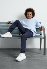 Helles blaugraues Sweatshirt, dunkelblaue Jogginghosen und weiße Turnschuhe, sitzen auf einer türkisfarbenen Bank mit einem glatten grauen Boden in einem minimalistisch gestalteten Hintergrund.