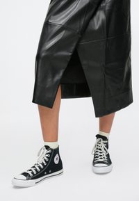 Benen met witte sokken en zwarte high-top Converse sneakers onder een zwarte leren midirok met zijsplit.