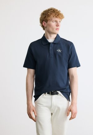 Noor punapea ja lokkis punaste juustega noormees kannab tumesinist Calvin Kleini polosärki ja valgeid pükse, vaadates paremale ühtlase tausta ees.