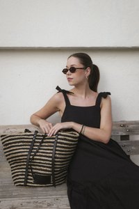 Straw tote bag with black and natural stripes, black leather accents, and two sturdy handles, placed next to a person wearing a black dress.