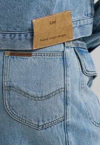 Φωτεινό μπλε τζιν με ορατή πίσω τσέπη και καφέ δερμάτινη ετικέτα ραμμένη με τα λογότυπα "Lee" και "Feng Chen WANG".