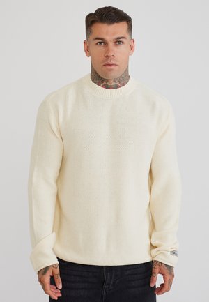 Mand med hals- og håndtatoveringer iført en cremet strikket sweater og sorte jeans, stående mod en ensfarvet lys baggrund.