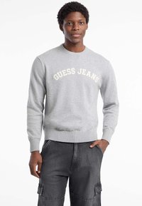 Suéter gris claro con "GUESS JEANS" en crema en el pecho, con cuello y puños acanalados, hecho de una suave tela de punto. Combinado con pantalones oscuros.