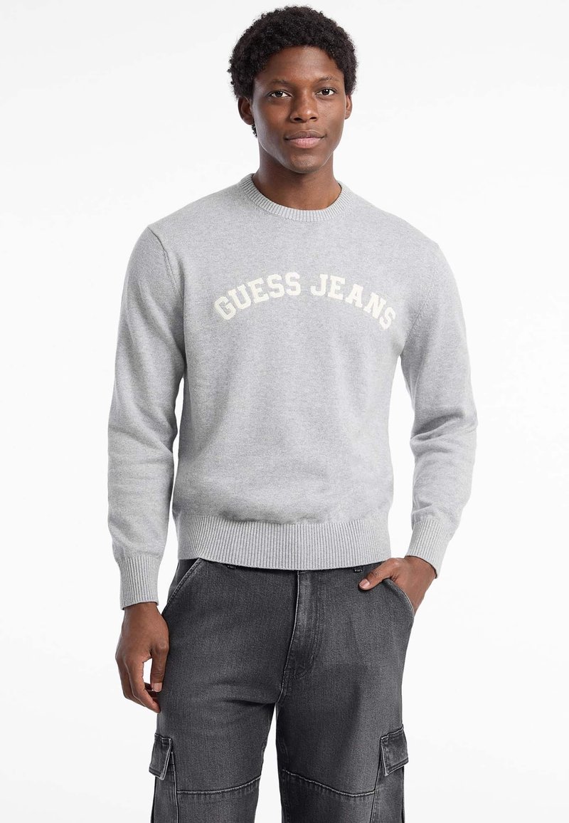Světle šedý svetr s nápisem "GUESS JEANS" v krémové barvě přes hrudník, s žebrovaným výstřihem a manžetami, vyrobený z měkké pleteniny. Doplněný o tmavé kalhoty.