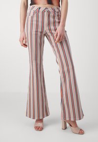 Pantalon évasé rayé en blanc, rouge et bleu. Il présente une taille haute, des poches avant et une texture de tissu lisse. Associé �à des talons beiges.