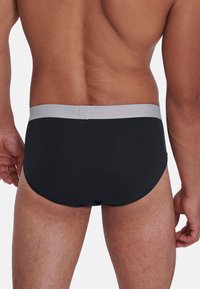 Uomo che indossa slip neri con cintura grigia chiara, visto da dietro, con tono della pelle visibile e peli sul corpo sulle gambe e sulla parte bassa della schiena.