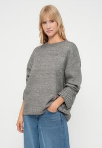 Sivi oversized pulover s rebrastim okruglim izrezom i manžetama. Rebrasti detalj na donjem rubu. Nositi s plavim širokim trapericama.