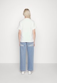 Witte katoenen t-shirt met korte mouwen, gecombineerd met lichtgewassen wijde jeans. Witte sneakers worden gedragen, weergegeven vanachter.