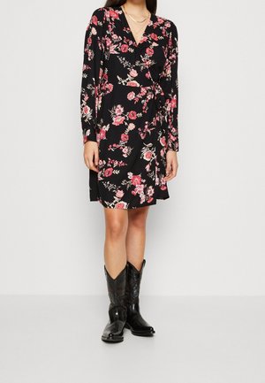 Vrouw die een zwarte bloemen wikkeljurk met lange mouwen en zwarte cowboylaarzen draagt, staand tegen een effen lichte achtergrond.