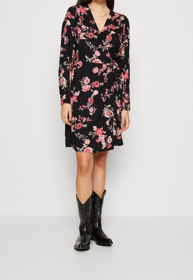 Femme portant une robe portefeuille noire à motif floral avec des manches longues et des bottes de cow-boy noires, debout devant un fond clair uni.