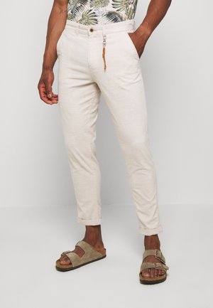 Pantalon en lin beige léger avec une coupe slim, doté d'une fermeture à bouton, de poches latérales et de bas roulés. Porté avec des sandales marron.