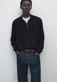 Young man stands against white wall wearing black long-sleeve zip collar shirt, white undershirt, blue jeans, and brown belt with hands in pockets.