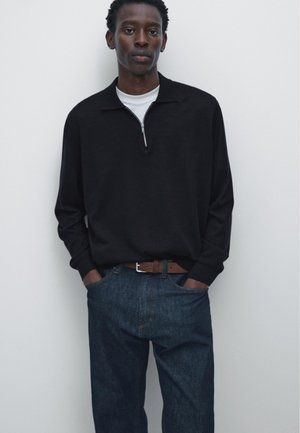 Young man stands against white wall wearing black long-sleeve zip collar shirt, white undershirt, blue jeans, and brown belt with hands in pockets.