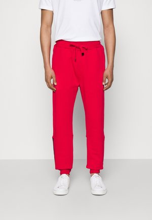 Homme portant un pantalon de jogging rouge vif avec taille à cordon, baskets blanches et un t-shirt blanc uni à manches courtes, debout devant un fond clair.