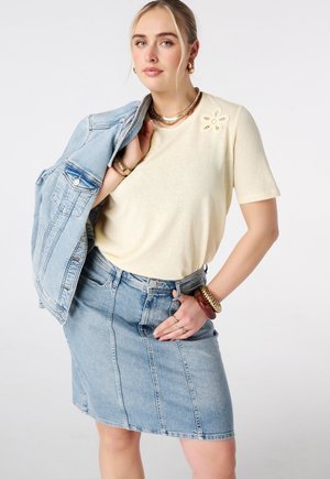 Jonge vrouw die een crèmekleurige top met korte mouwen en bloemmotief draagt, een lichte denim rok, en een spijkerjasje over haar schouder hangt.