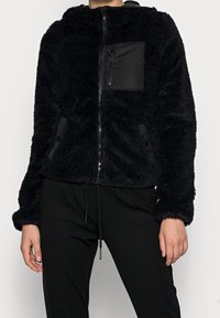 Giacca in pile nera con zip frontale, cappuccio, tessuto peloso testurizzato e tasca sul petto; presenta accenti di pannelli neri lisci a contrasto.