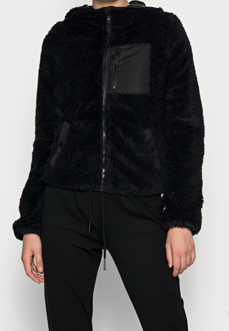 Giacca in pile nera con zip frontale, cappuccio, tessuto peloso testurizzato e tasca sul petto; presenta accenti di pannelli neri lisci a contrasto.