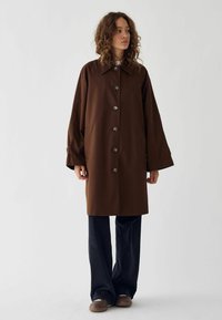 Bruine, oversized jas met een grote kraag, knopen aan de voorkant en zijzakken. G styled met een wijdvallende donkere spijkerbroek en bruine schoenen. Vlotte stofstructuur.