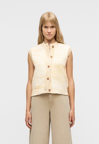 Γυναίκα με ίσια ξανθά μαλλιά, φορώντας αμάνικο πλεκτό γιλέκο σε μπεζ και λευκό με ξύλινα κουμπιά και ανοιχτό καφέ παντελόνι με ευρύ πόδι.