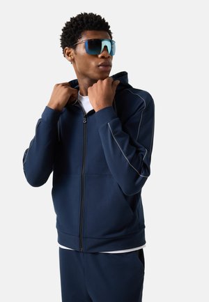 Młody mężczyzna poprawiający kaptur granatowej kurtki na zamek, noszący ciemnoniebieskie refleksyjne okulary przeciwsłoneczne, białą koszulę i pasujące granatowe spodnie, patrzy w prawo.