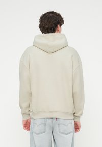 Ανοιχτό μπεζ φούτερ με μεγάλο κουκούλι, χαλαρούς ώμους, ριμπ μανσέτες και τελείωμα, φορεμένο πάνω από ανοιχτό μπλε τζιν.