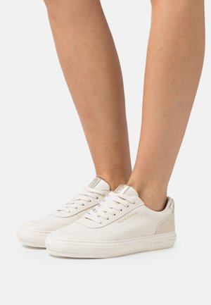 Baskets basses blanches à lacets avec la marque dorée "Marc O'Polo", portées pieds nus sur un fond uni.