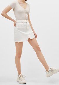 Biała spódnica z denimu z postrzępionym dołem, z przednim rozcięciem i jasnym przeszyciem. Połączona z beżową prążkowaną bluzką i białymi tenisówkami.