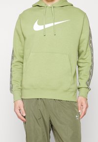 Grön hoodie med en stor vit Nike-logotyp, kenguruficka och kontrasterande svarta vertikala ränder längs ärmarna. Mjuk tyg, standard passform.