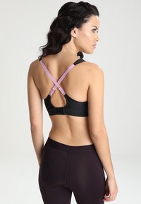 Svart sportsbh med krysset rosa stropper, justerbar bakre krok-lukking og pustevennlig stoff. Modellen har på seg mørke tights.