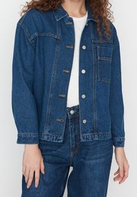 Tummansininen farkkutakki, jossa on napitettava etuosa, rintatasku ja pitkät hihat. Rakenteellinen kangas, jossa näkyvät ompelusaumat.