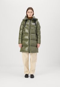 Gevoerde parka in olijfgroen met glanzende afwerking, lange silhouet, verstelbare capuchon en zwarte banden; gecombineerd met crèmekleurige broeken en zwarte schoenen.