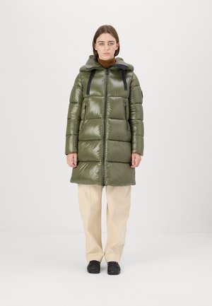 Gevoerde parka in olijfgroen met glanzende afwerking, lange silhouet, verstelbare capuchon en zwarte banden; gecombineerd met crèmekleurige broeken en zwarte schoenen.