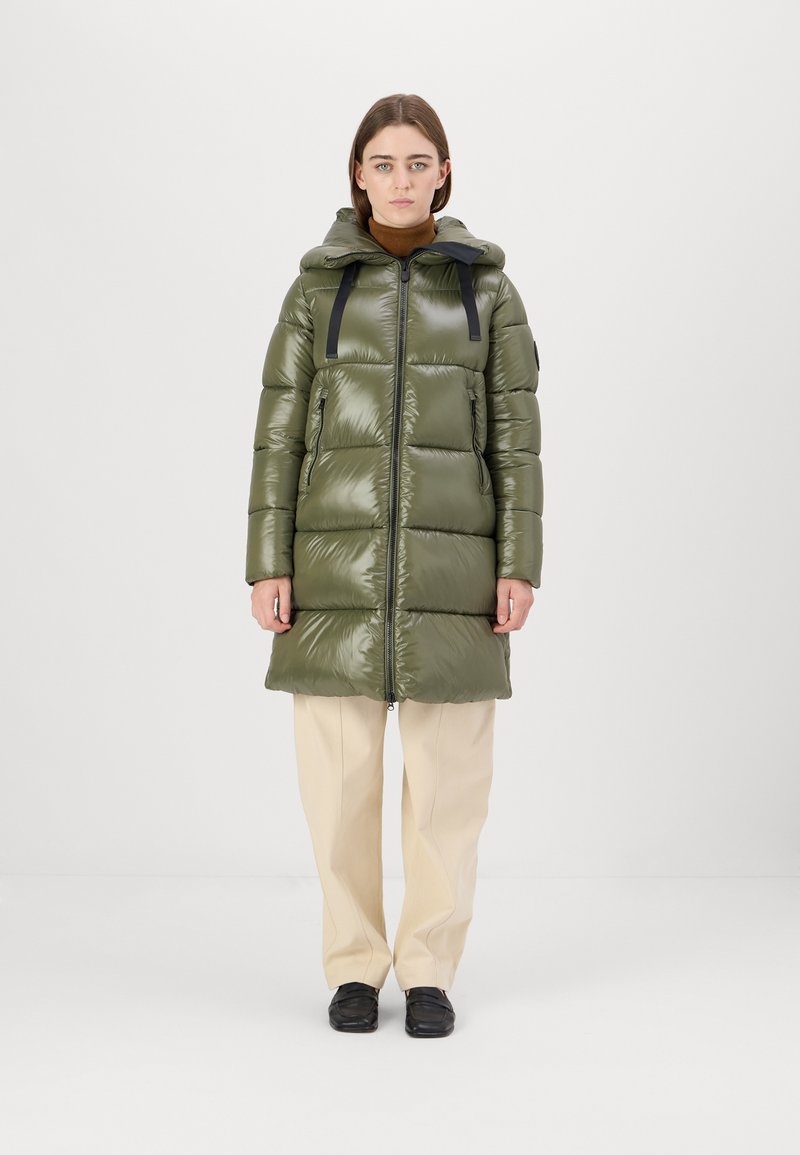 Gevoerde parka in olijfgroen met glanzende afwerking, lange silhouet, verstelbare capuchon en zwarte banden; gecombineerd met crèmekleurige broeken en zwarte schoenen.