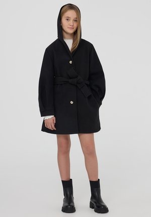 Giovane donna che indossa un cappotto nero con cintura e cappuccio, una maglietta bianca a maniche lunghe e stivaletti neri, in piedi contro uno sfondo neutro.