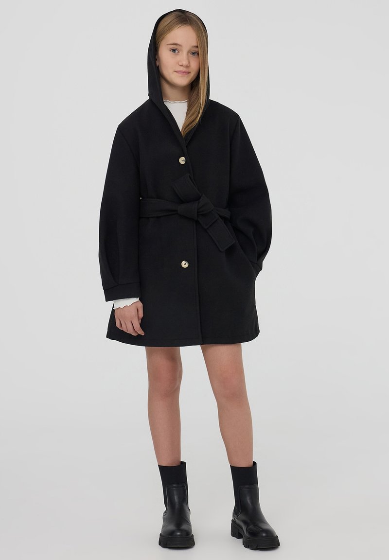 Giovane donna che indossa un cappotto nero con cintura e cappuccio, una maglietta bianca a maniche lunghe e stivaletti neri, in piedi contro uno sfondo neutro.