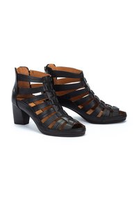 Botas de tobillo de cuero negro, con punta abierta y un diseño trenzado, tacón apilado, cremallera lateral y forro interior beige. Acabado texturizado y correas ajustables.