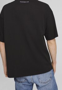 Camiseta de algodón negra con un corte relajado, mangas cortas y un pequeño logo de "Porsche" impreso en blanco en la parte posterior del cuello. Llevada con pantalones vaqueros azules.