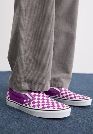 Scarpe Vans slip-on a scacchi viola e bianchi, con superficie liscia e bordo nero a contrasto, abbinate a pantaloni in corduroy grigi.